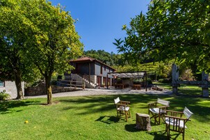 Una casa rústica con una escalera de piedra que conduce a una terraza de madera, rodeada de abundante vegetación y un cielo azul despejado.
