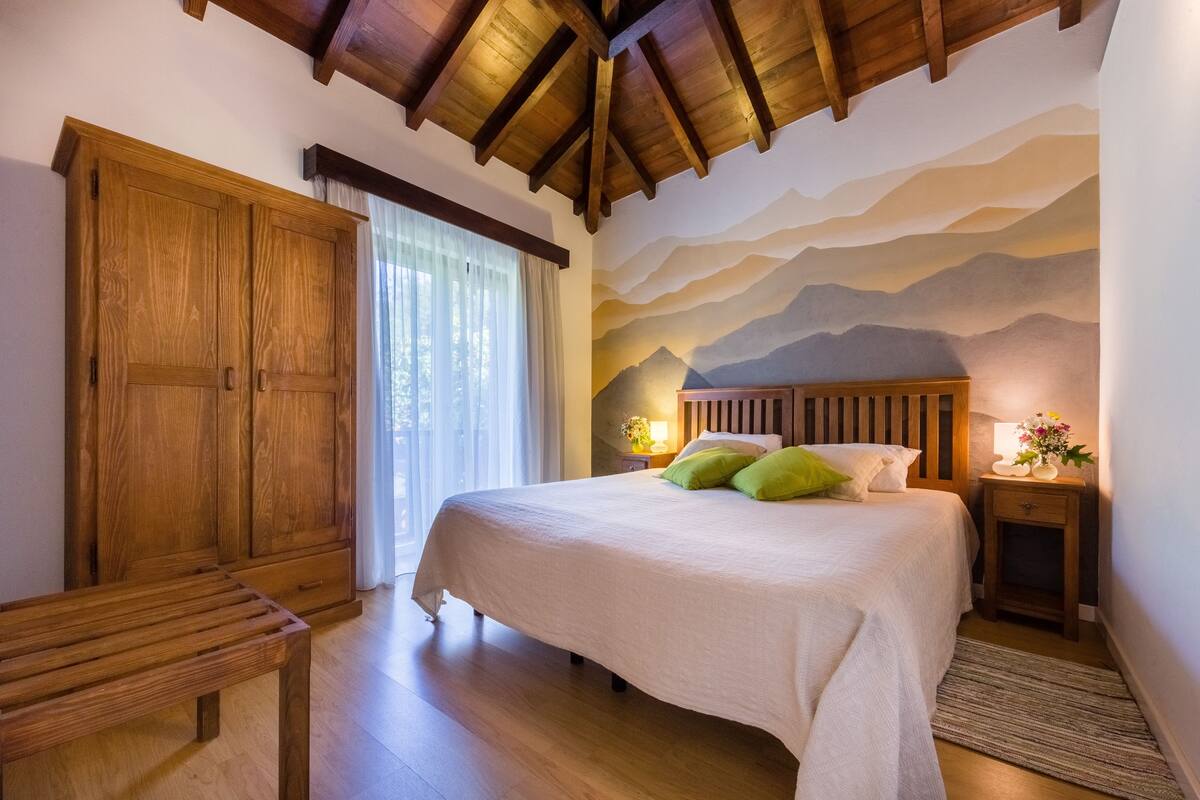 A bedroom with a wooden bed, a large wardrobe, and a mountain mural on the wall.