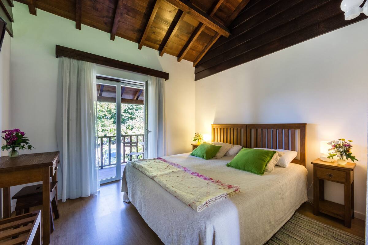 A bedroom with a large bed, wooden headboard, bedside tables with lamps, a window with curtains, and a view of greenery outside.