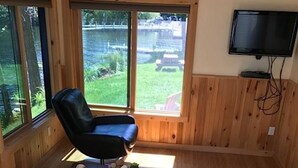 A cozy living room with a leather chair, a wooden coffee table with decorative items, a white sofa, and large windows offering a view of a grassy yard. - Relax on Crescent Lake  #2 (Wolfeboro)