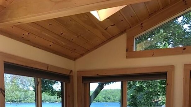 A wooden interior with a round table and four chairs, large windows overlooking a body of water, and a view of a dock with a covered area. - Relax on Crescent Lake  #2 (Wolfeboro)