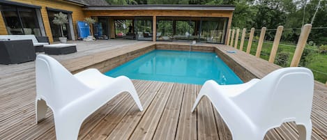Une terrasse en bois avec une piscine, des chaises longues blanches et une maison moderne en arrière-plan.