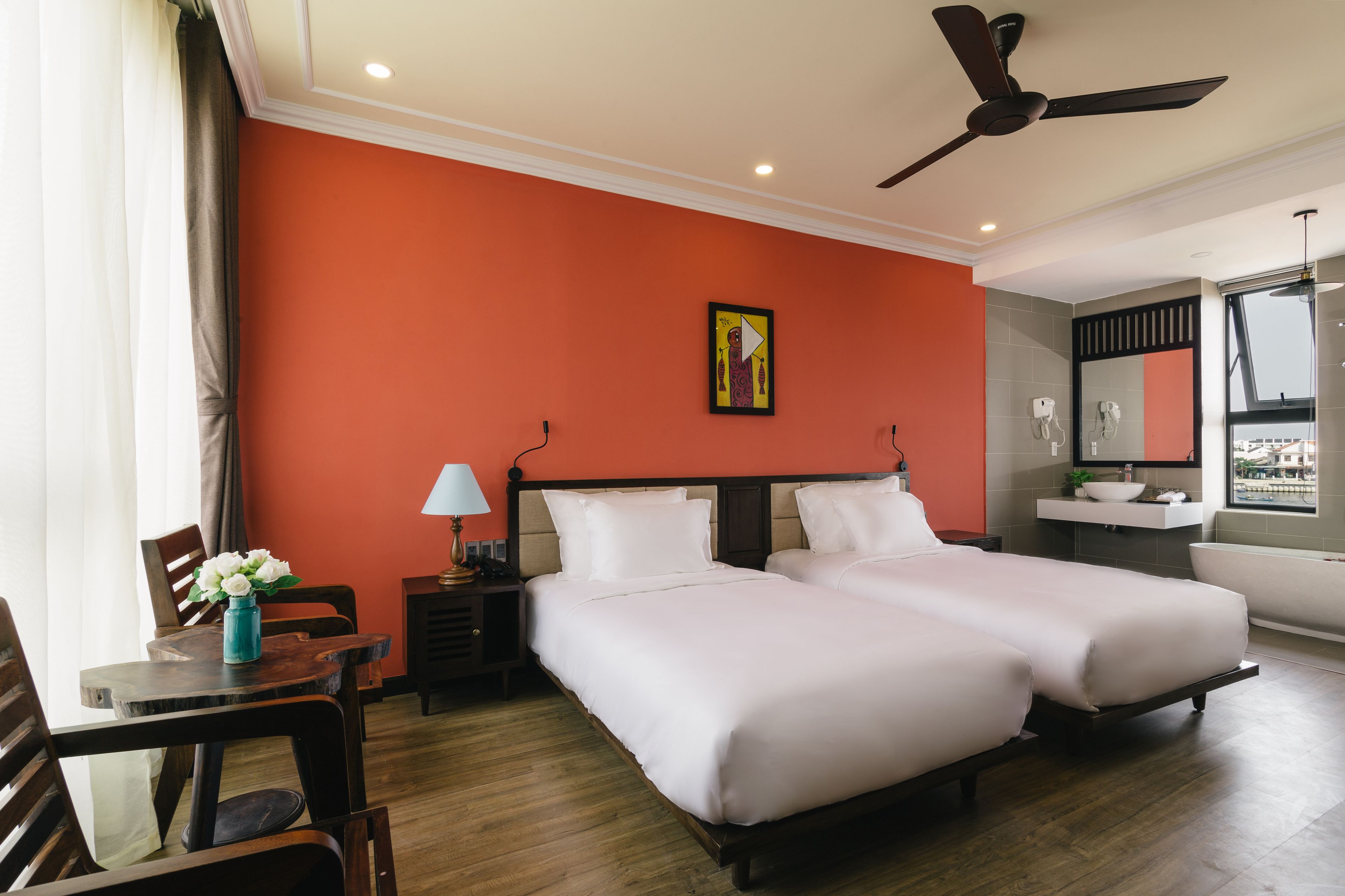 Une chambre d’hôtel avec deux lits, un bureau, une chaise, une table de chevet, une lampe, un ventilateur de plafond et une fenêtre donnant sur un beau paysage.