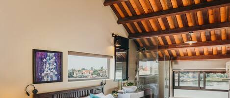 A bedroom with a wooden ceiling, a bed with white bedding, a nightstand with a lamp, a bathroom with a bathtub, and a window with a view of buildings.