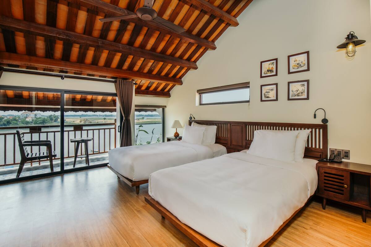 A hotel room with two beds, a balcony with a view, wooden flooring, and a ceiling fan.