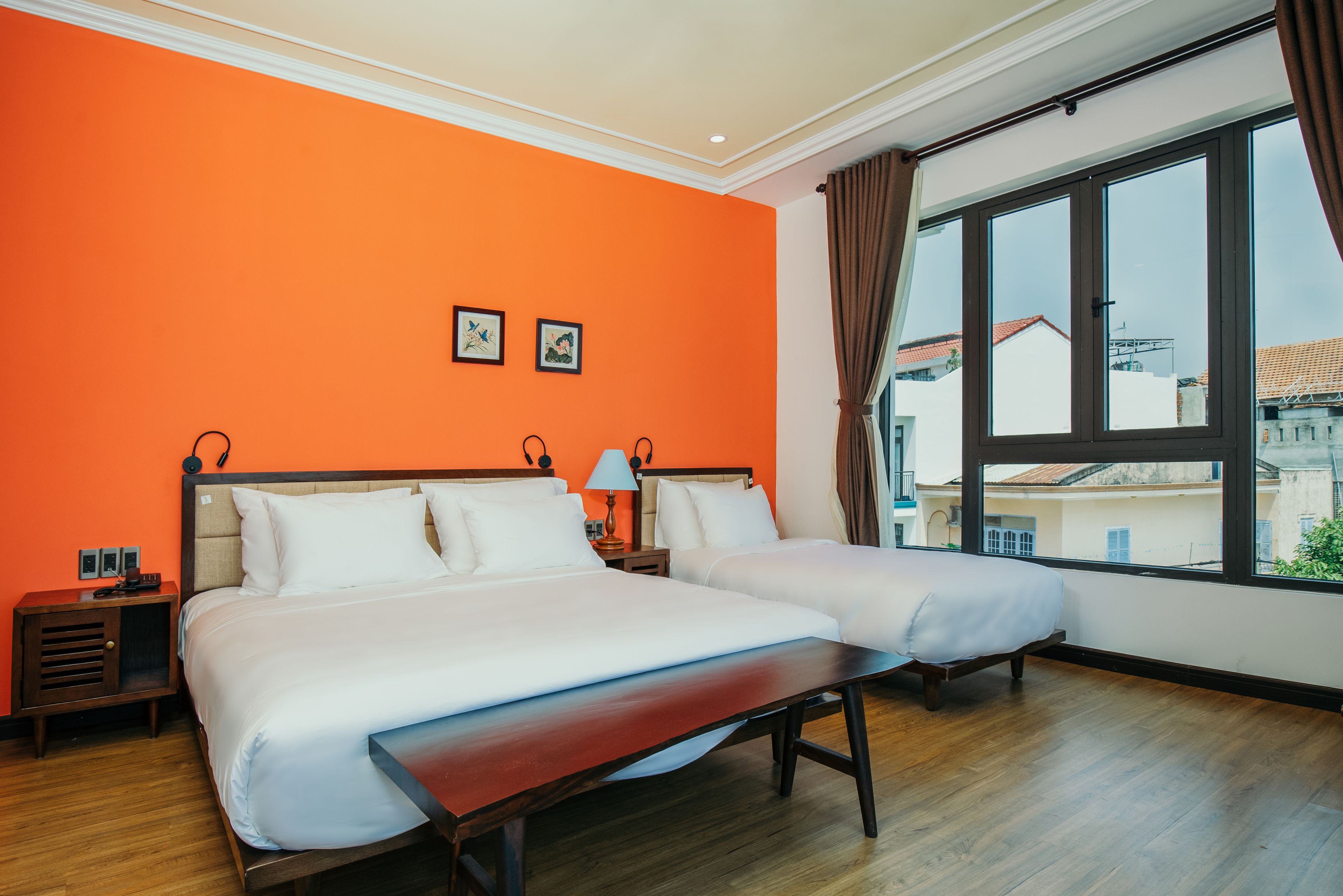 A hotel room with two beds, a wooden bench, a nightstand with a lamp, and a large window offering a city view.