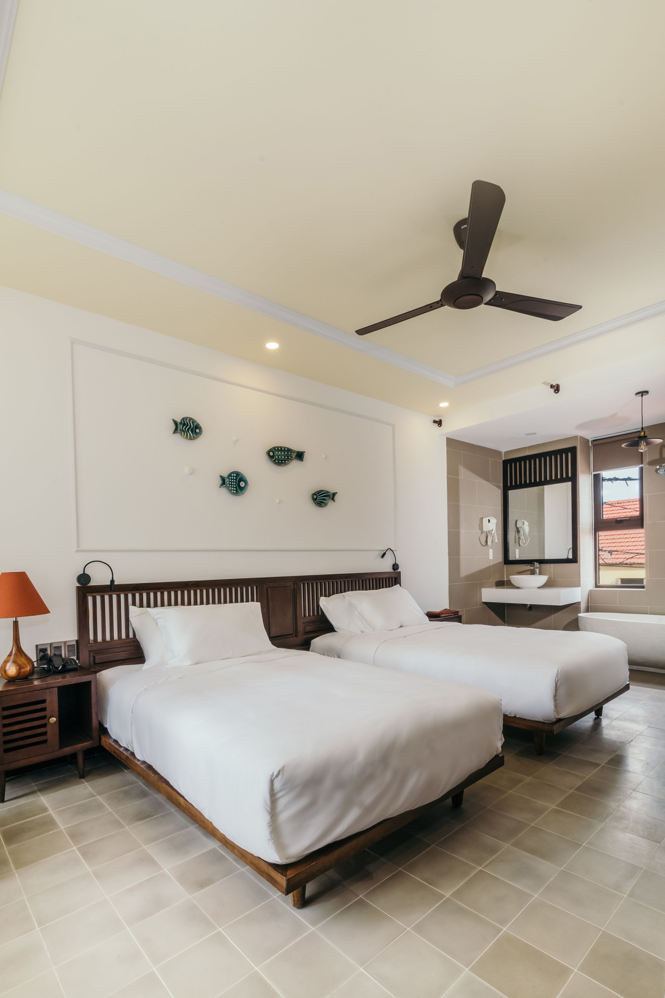 Habitación de hotel con dos camas, un ventilador de techo, un adorno de pez en la pared, una tocador con espejo y una ventana con vistas.
