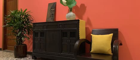 A room with a patterned floor, a wooden door, a black cabinet with a Buddha statue, a vase with flowers, and a chair with a yellow cushion.