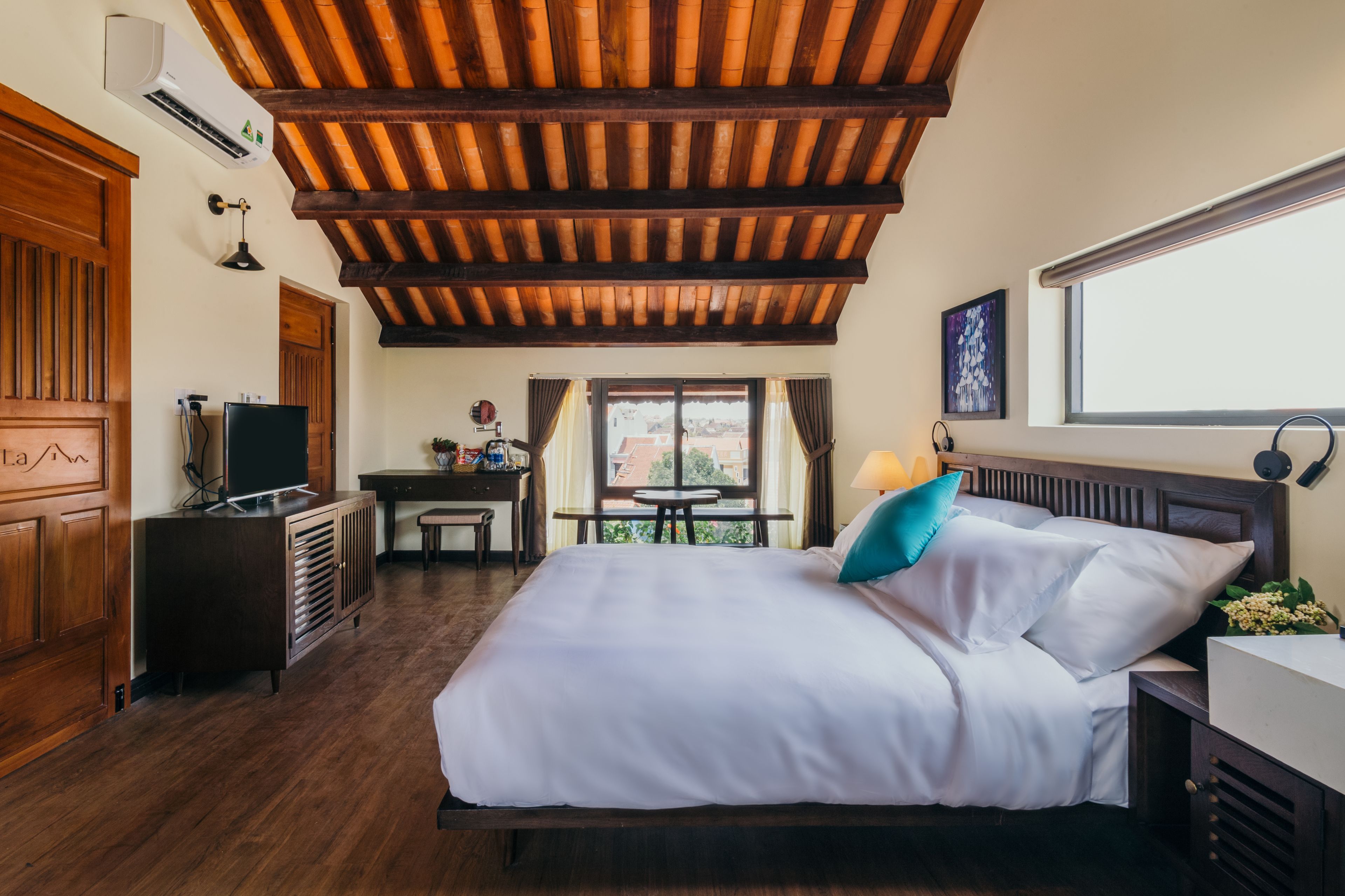 Een hotelkamer met een houten plafond, een bed met wit beddengoed, een bureau met een computer en een raam met gordijnen.