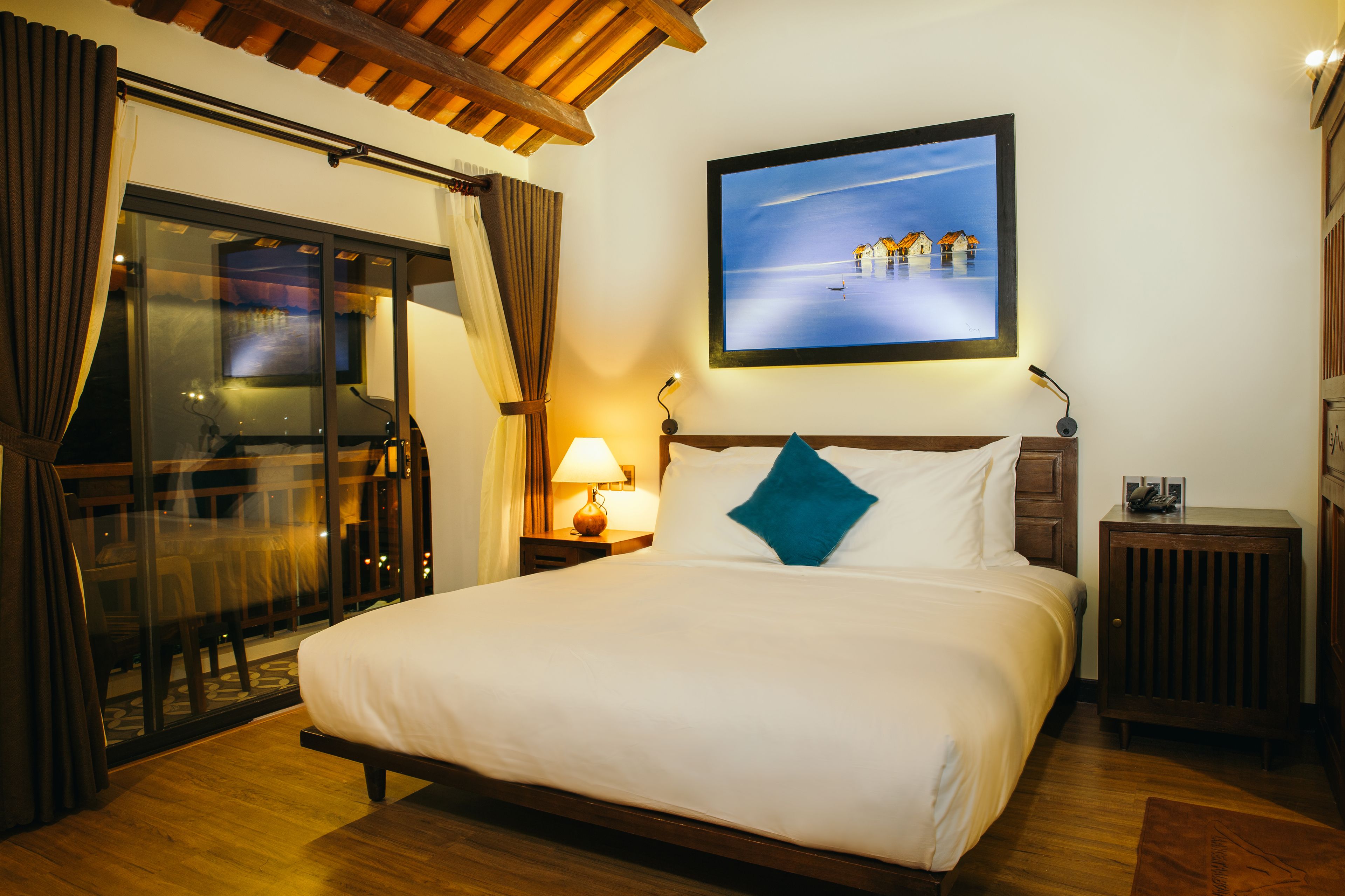 Una camera d'albergo con un letto grande, una porta vetrata che dà su un balcone, un quadro appeso al muro e un comodino con una lampada.
