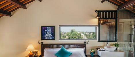 A bedroom with a wooden ceiling, a bed with white bedding, a window with a view, a bedside table with a lamp, and a bathroom with a glass shower.
