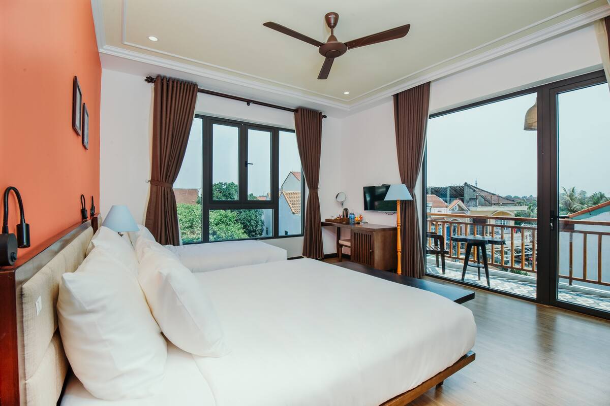 A hotel room with a large bed, a desk with a chair, a TV, and a balcony with a view of the city.