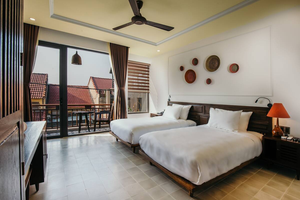A hotel room with two beds, a ceiling fan, a desk, a chair, a balcony with a view of buildings, and a wall with decorative plates.
