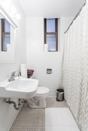 A bathroom with a white sink, toilet, and shower curtain. There are two windows, a trash can, and a towel rack. - Live Like a Local - Downtown 3 BDR Gem (Chicago)