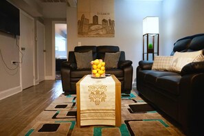 A living room with two black leather sofas, a coffee table with a decorative tablecloth, a wall-mounted cityscape artwork, and a floor lamp. - The Karlov Connection - Ohare Wrigley Gateway (Chicago)
