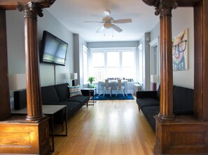 A modern living room with a wooden floor, a black sofa, a dining table with chairs, and a ceiling fan. - Classic Lincoln Park Charmer - 3bdr Steps to Train (Chicago)