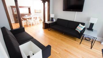 A modern living room with a black sofa, a white armchair, and a wooden floor. A flat-screen TV is mounted on the wall, and a small table with a lamp is visible.