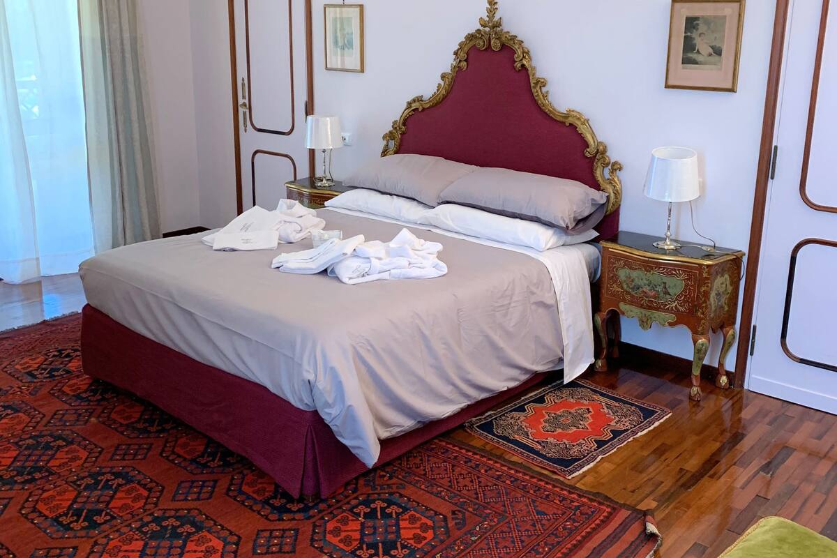 A neatly made bed with a red headboard, two bedside tables with lamps, a patterned rug, and a green ottoman.