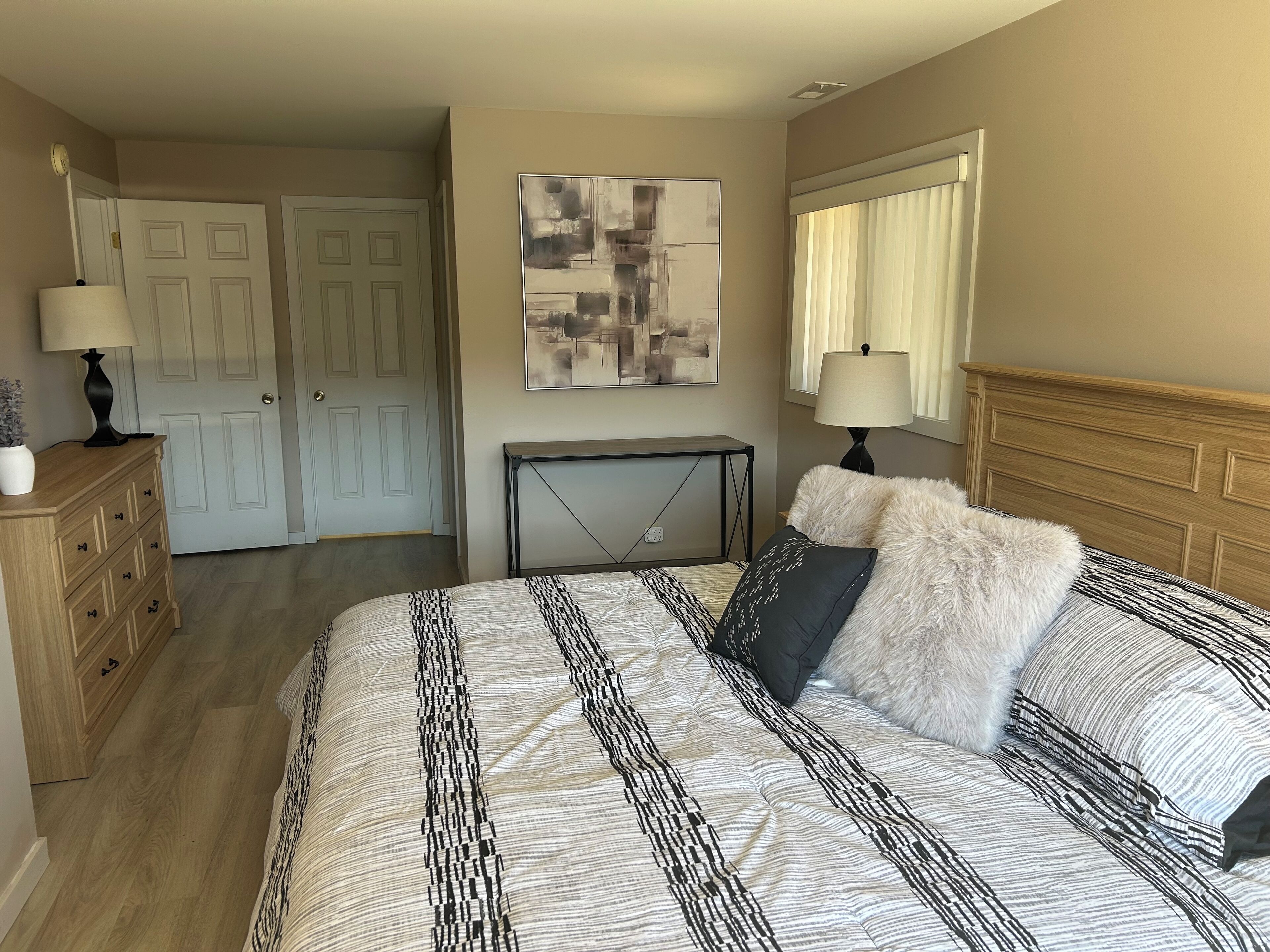 A bedroom with a large bed, a wooden dresser, a console table, and a painting on the wall.