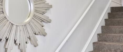 Un escalier avec une rampe blanche et des marches recouvertes de moquette, une console blanche avec des tiroirs et un miroir en forme de soleil accroché au mur.