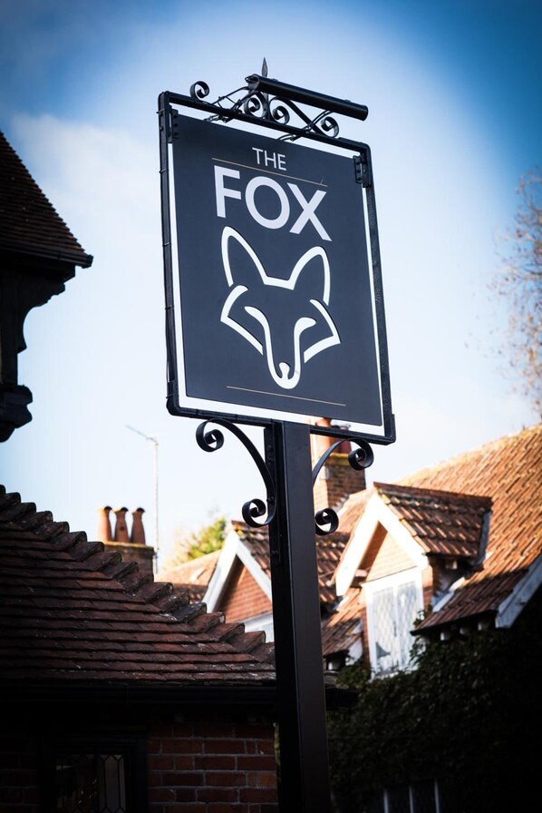 Señal del pub 'The Fox' con un logotipo estilizado de cabeza de zorro, montada en un poste de metal con detalles decorativos en forma de volutas.