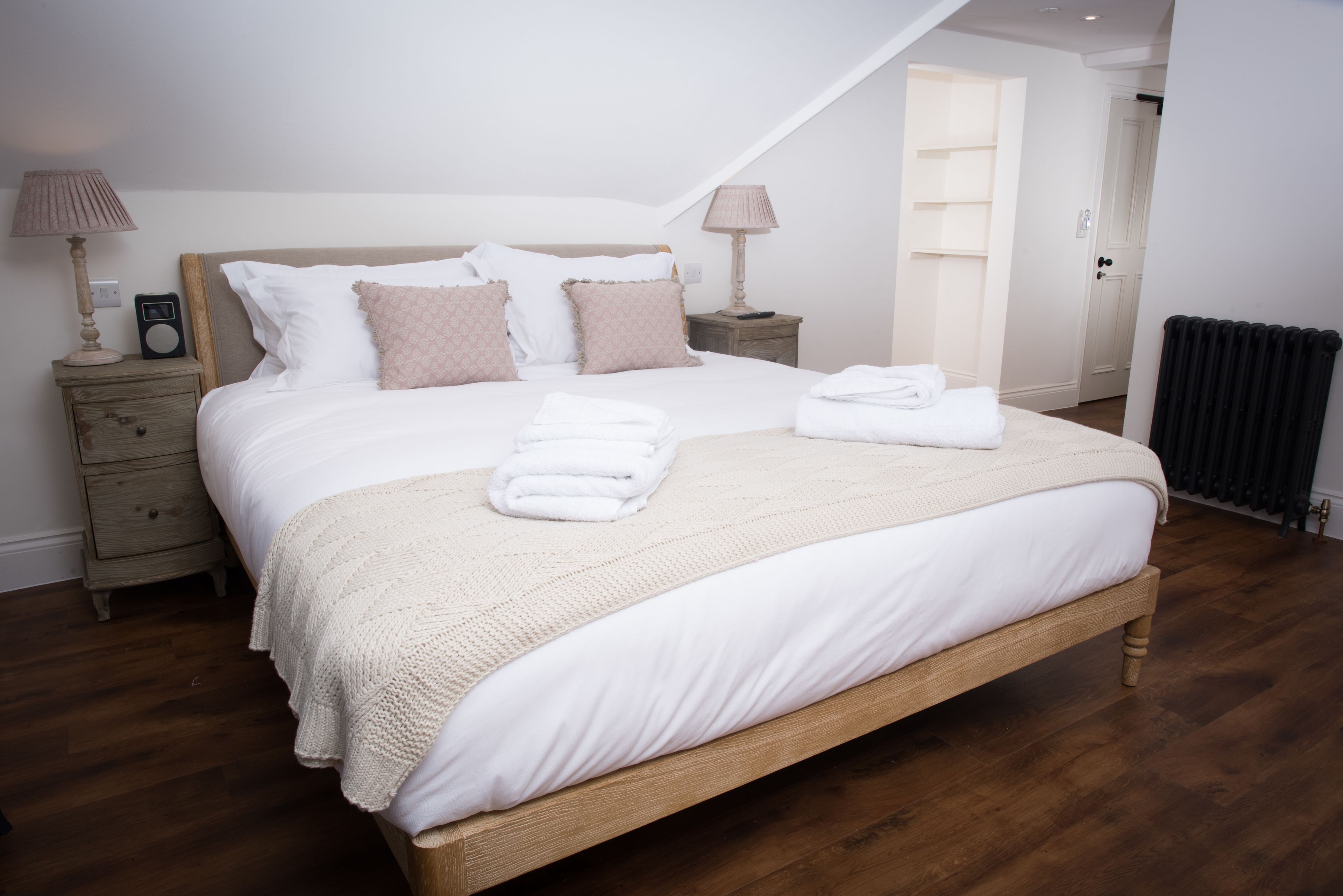 Una cama bien hecha con sábanas blancas y una manta color beige, dos mesitas de noche con lámparas y piso de madera.