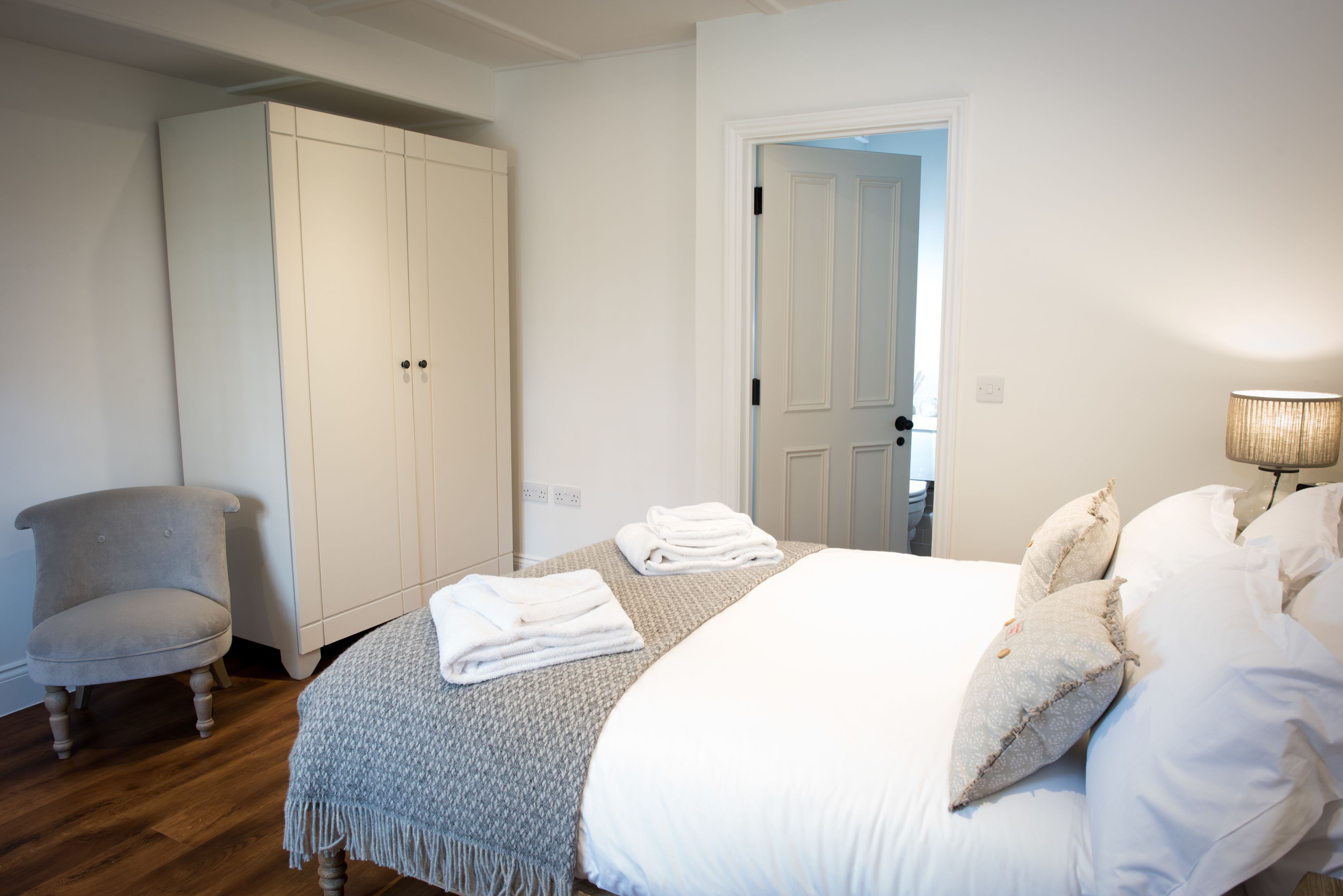 A bedroom with a large wardrobe, a chair, a neatly made bed with towels, and a lamp.
