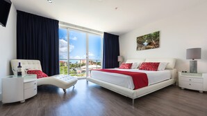A modern hotel room with a large bed, a seating area, a desk, and a large window offering a city view. - Las Islas Aruba (Noord)