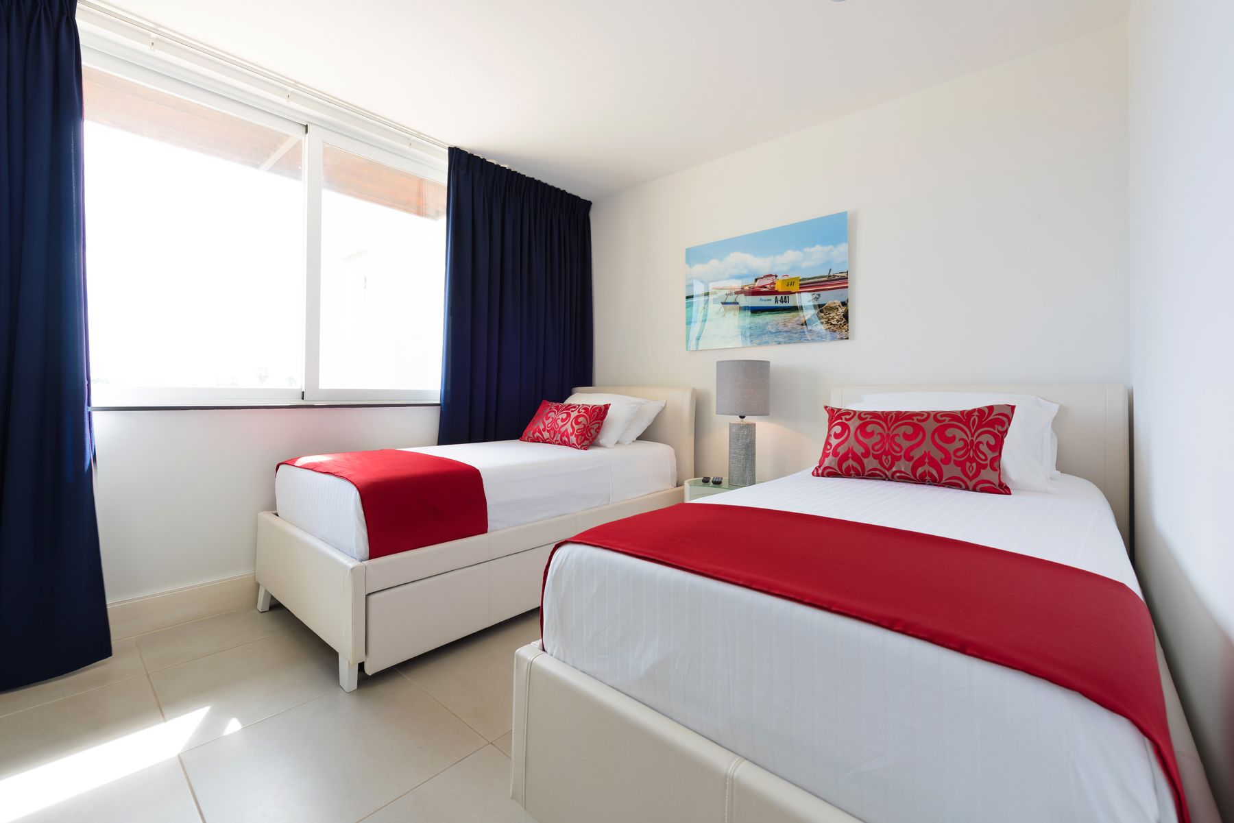 Une chambre d’hôtel avec deux lits, chacun recouvert d’une couverture rouge et blanche, une table de chevet avec une lampe et un tableau représentant un bateau accroché au mur.