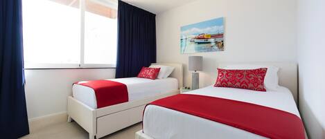 Une chambre d’hôtel avec deux lits, chacun recouvert d’une couverture rouge et blanche, une table de chevet avec une lampe et un tableau représentant un bateau accroché au mur.