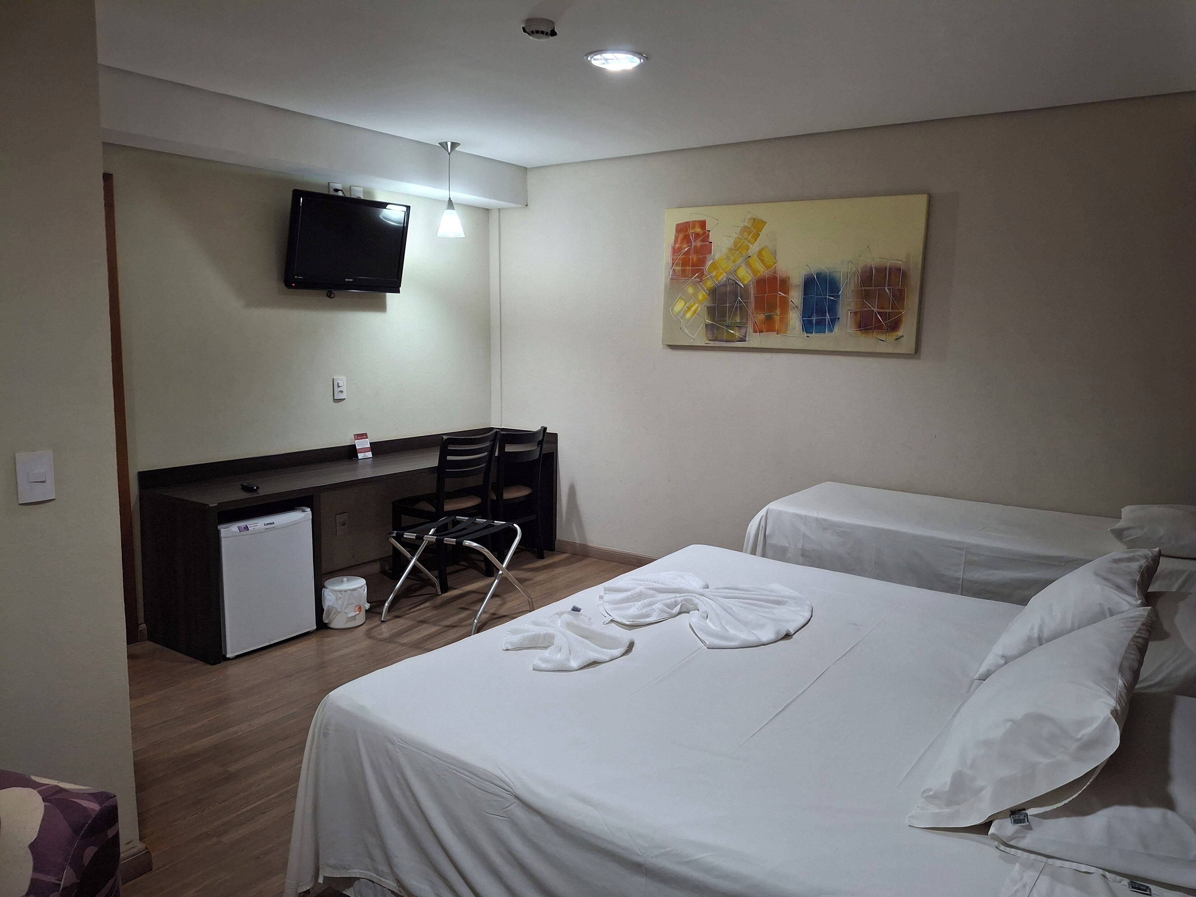 A hotel room with a bed, a desk, a chair, a television, and a painting on the wall.