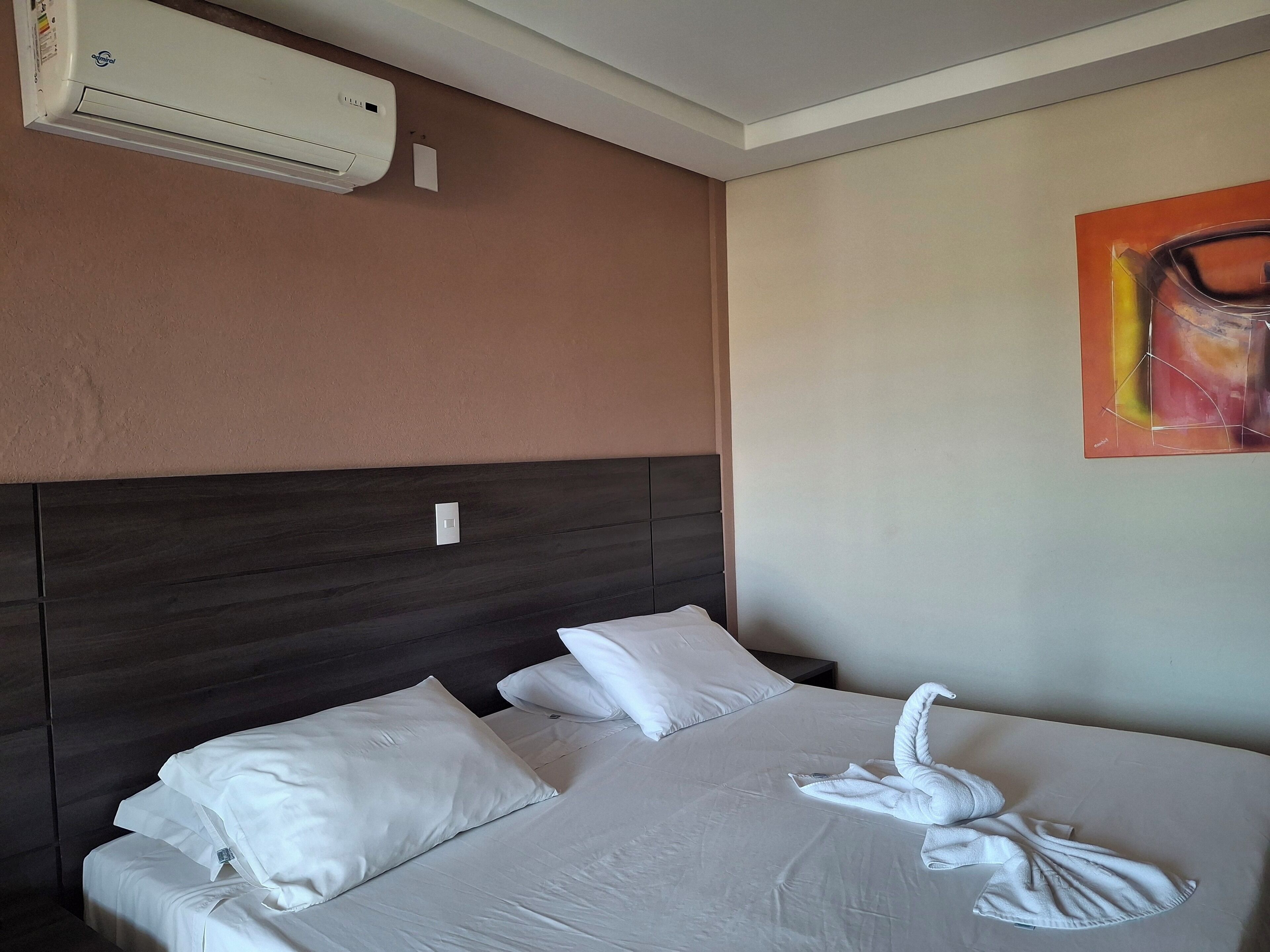 Habitación de hotel con cama, ropa de cama blanca, aire acondicionado de pared, un cuadro y un cabecero de madera oscura.