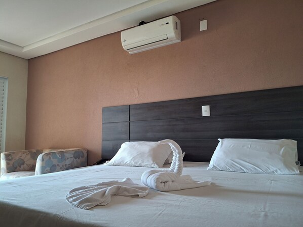 Una habitación de hotel con una cama, cabecera oscura, unidad de aire acondicionado, interruptor de luz en la pared y un sillón estampado.