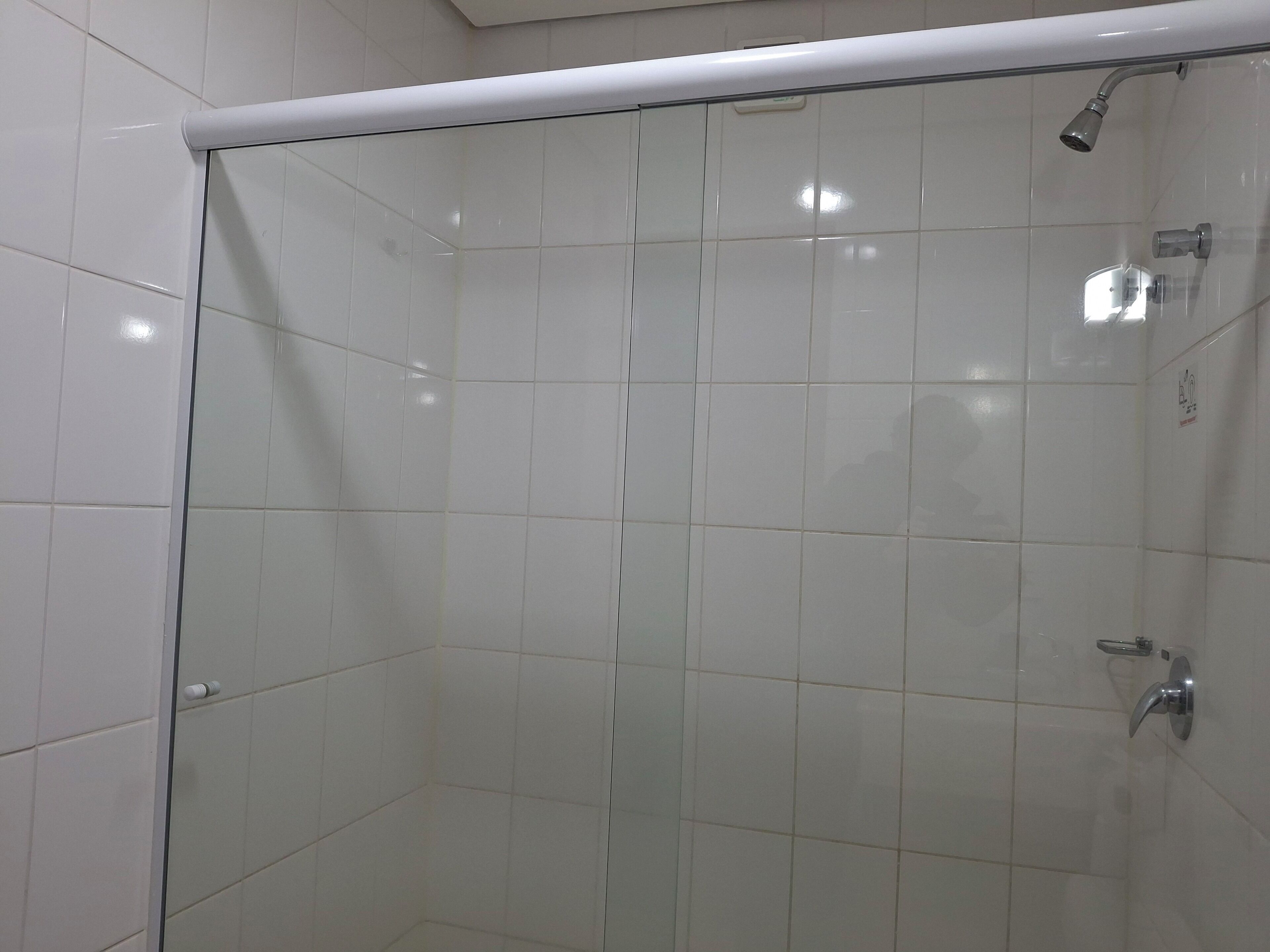 Eine Duschkabine mit Glas, weißen Fliesen, einer Handbrause und einem am Wand montierten Wasserhahn.