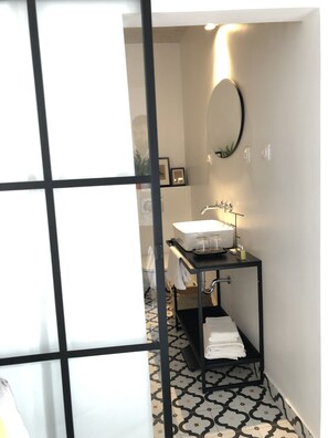 A bathroom with a black metal vanity, a white sink, and a mirror. There is a towel rack with folded towels and a small shelf with toiletries. The floor has patterned tiles.