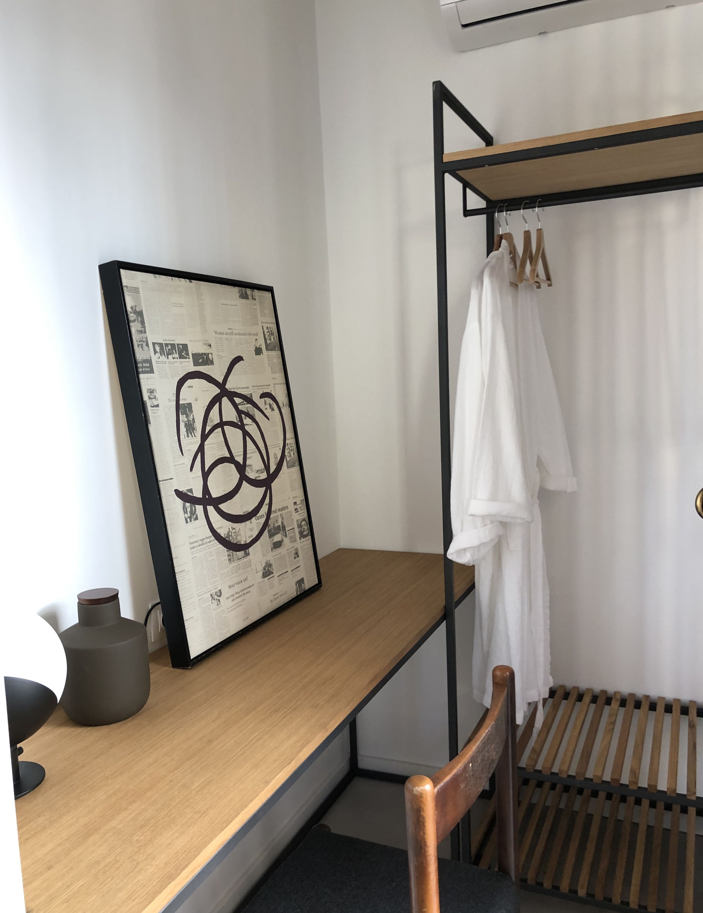 Une chambre moderne avec un bureau en bois, une œuvre d’art abstraite encadrée, une robe blanche posée sur un porte-manteau et une chaise noire.