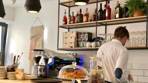 A bar area with a coffee machine, a glass display case with pastries, and shelves with bottles and glasses. - Casa Figa 39 (Zagreb)