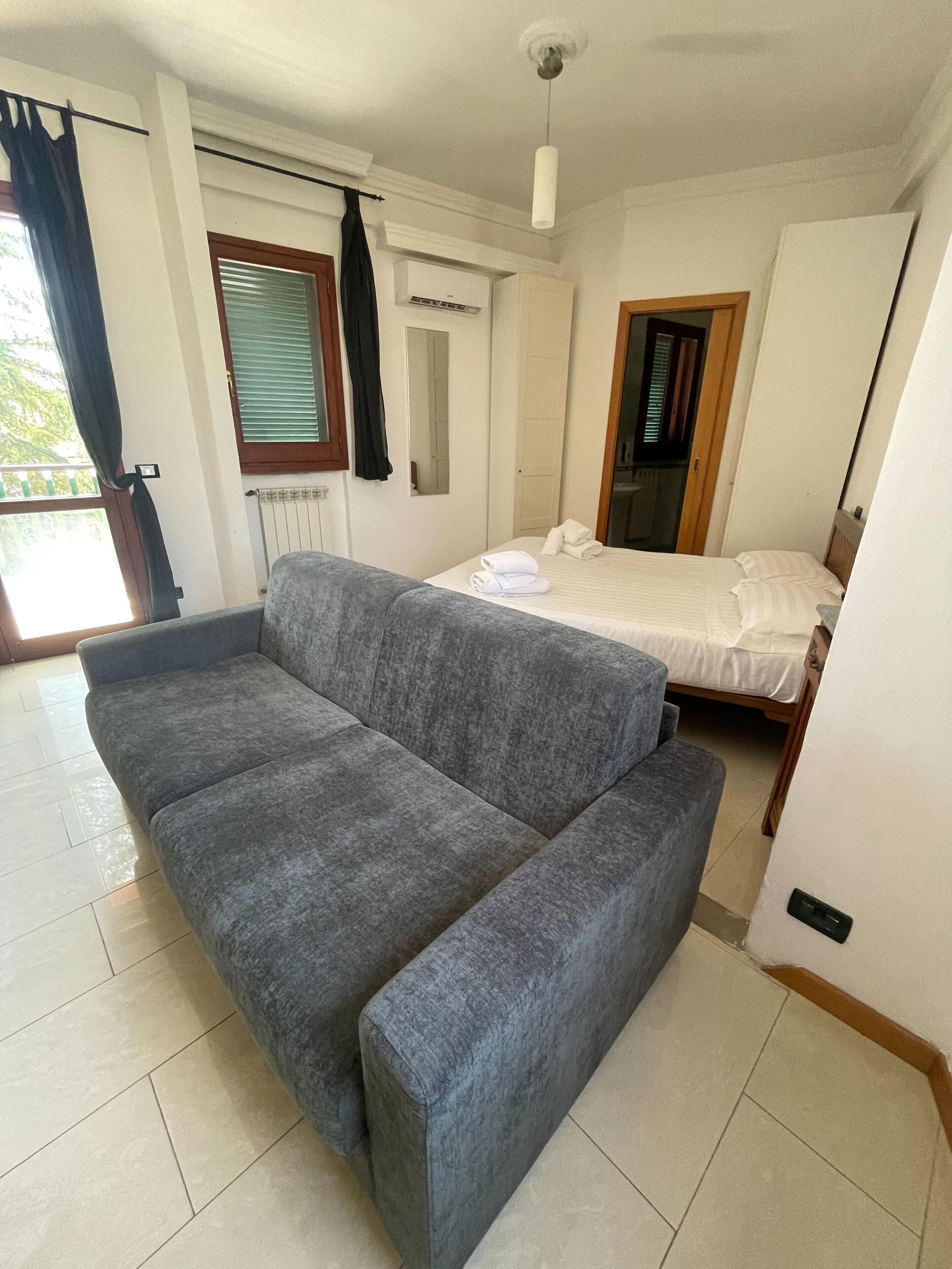 A room with a grey sofa, a bed with white bedding, a radiator, and a window with blinds.