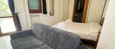 A room with a grey sofa, a bed with white bedding, a radiator, and a window with blinds.