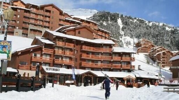 Une station de ski enneigée, avec des bâtiments en bois, dont un hôtel doté de nombreux balcons et d’une enseigne indiquant « SUPERSKIT ».