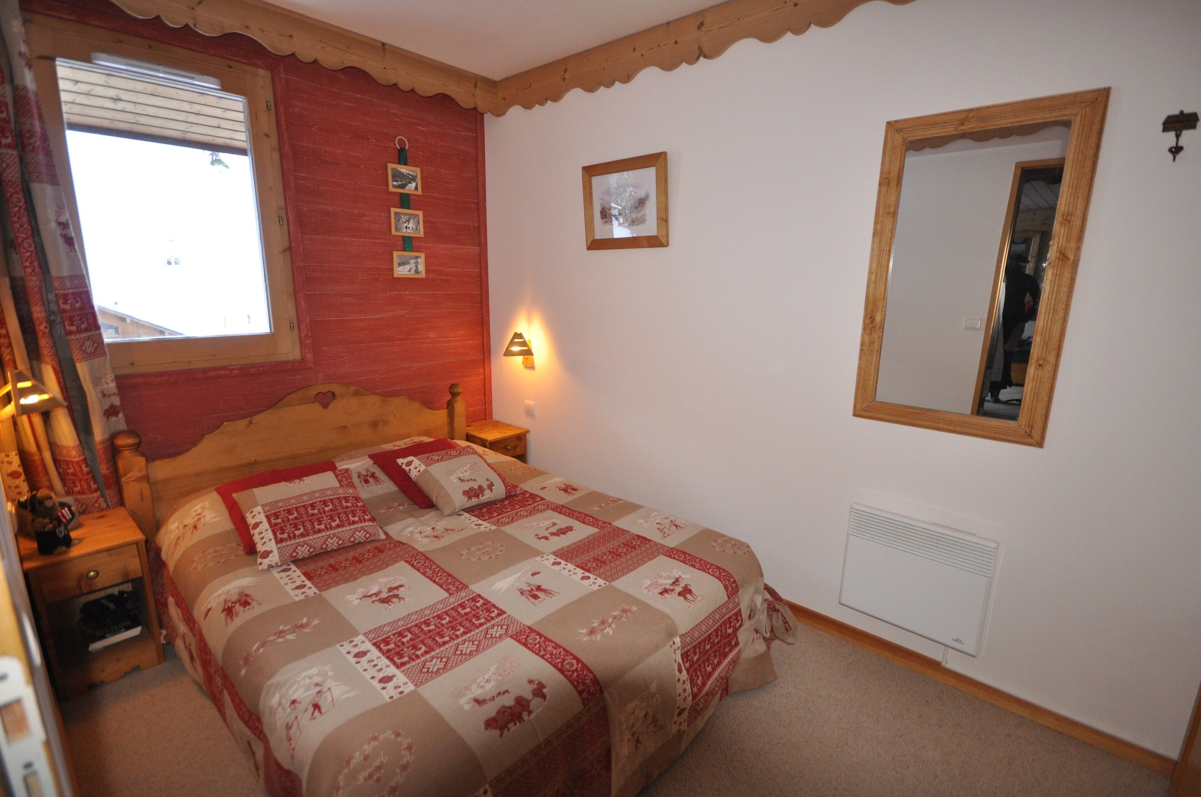 A bedroom with a wooden bed, a nightstand, a window with blinds, a wall-mounted mirror, and a small picture frame.