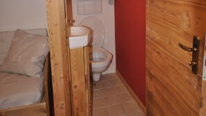 Une petite pièce compacte avec un plafond en bois, un mur d’accent rouge, une porte en bois, un lit, des toilettes et un petit lavabo.