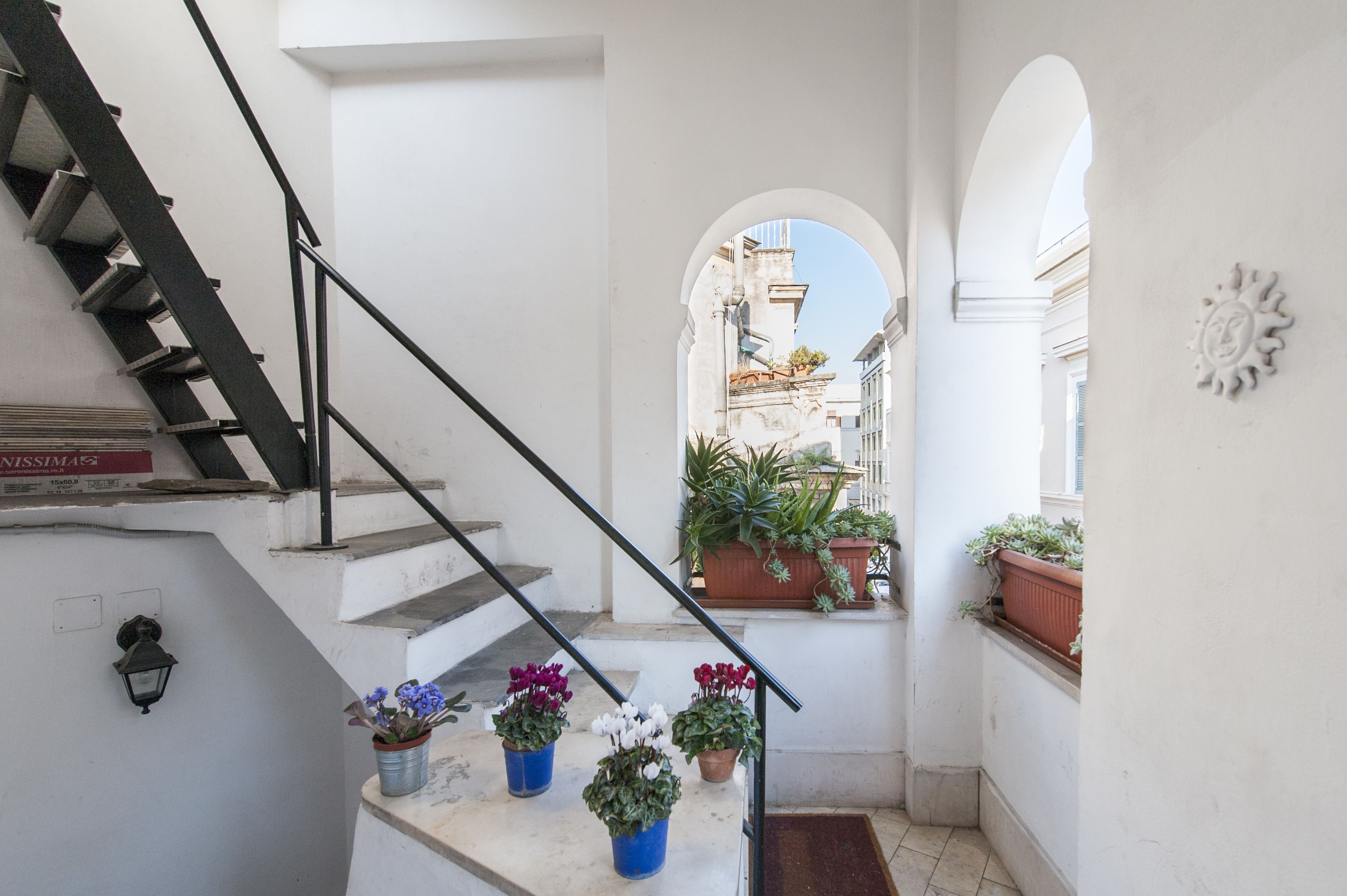 A narrow stairway with a metal railing, potted plants, and a view of buildings through an archway.