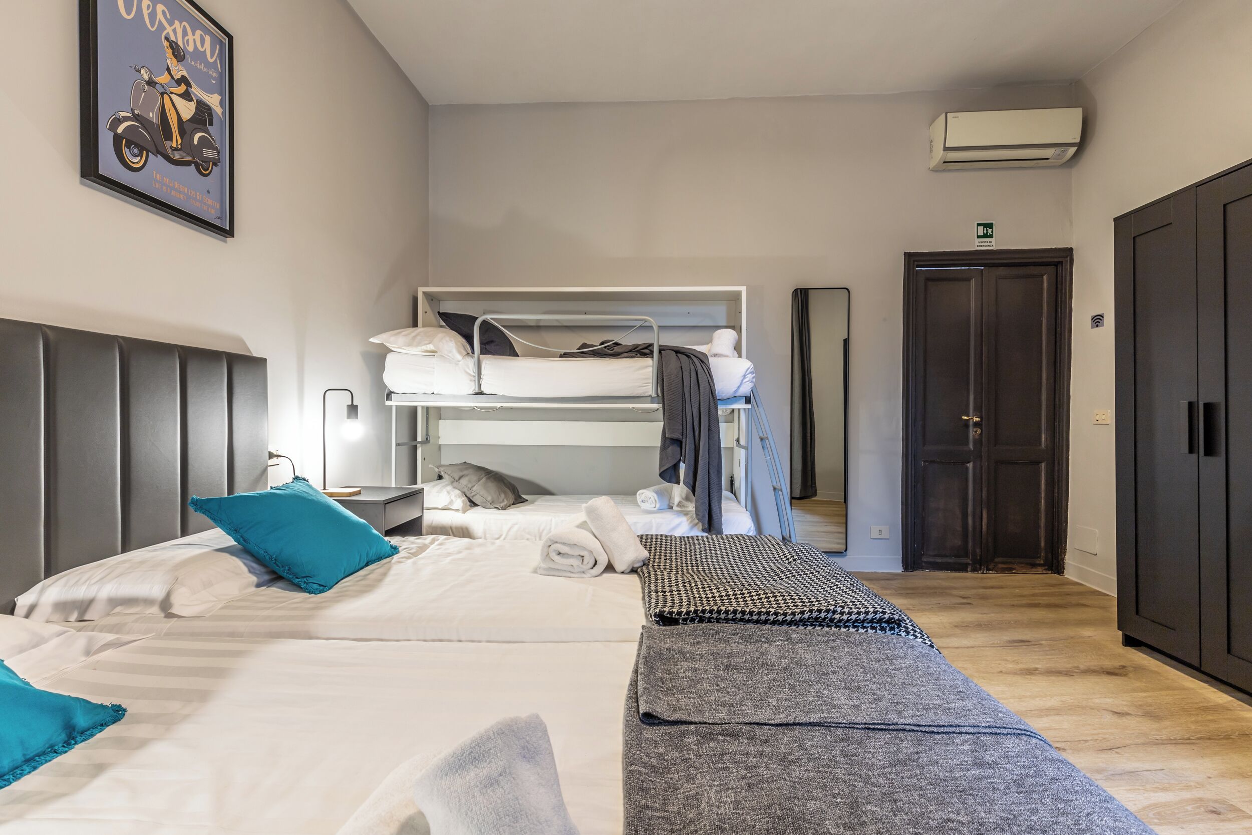 Hotelkamer met een stapelbed, een eenpersoonsbed, een nachtkastje, een wandairconditioner en een deur met een vluchtroutebord.
