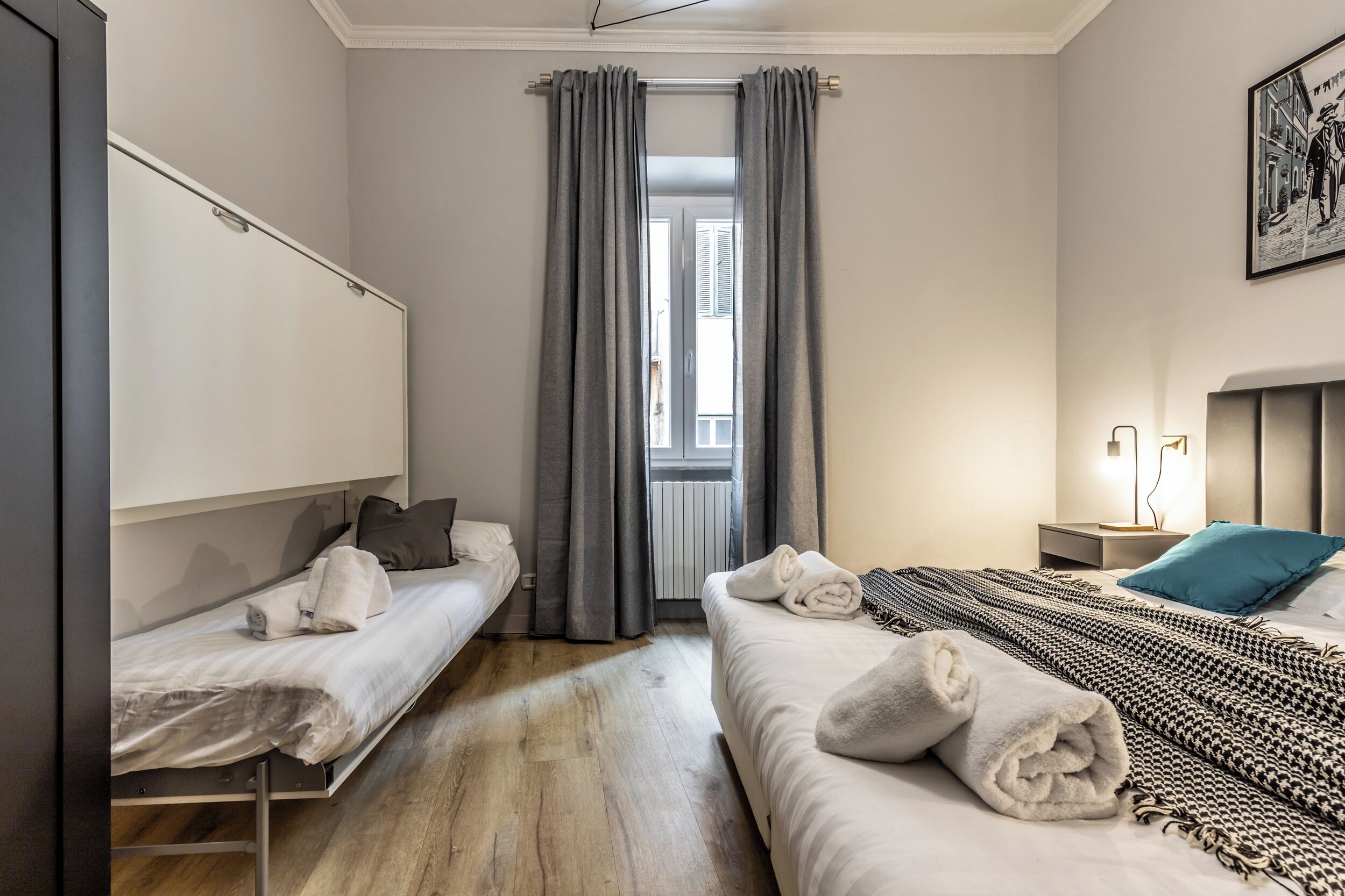 A bedroom with two single beds, a large window with grey curtains, a bedside table with a lamp, and a framed picture on the wall.
