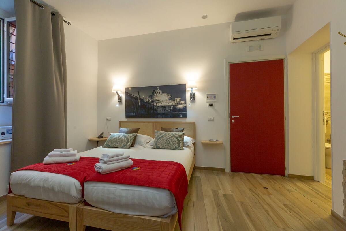 Un lit double avec une couverture rouge, une table de chevet, une lampe fixée au mur et une porte rouge.