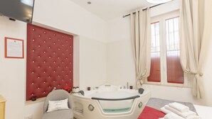 A modern bathroom with a round jacuzzi, a red tufted headboard, a window with red blinds, and a bed with neatly folded towels. - Flatinrome Trastevere Deluxe Rooms (Rome)