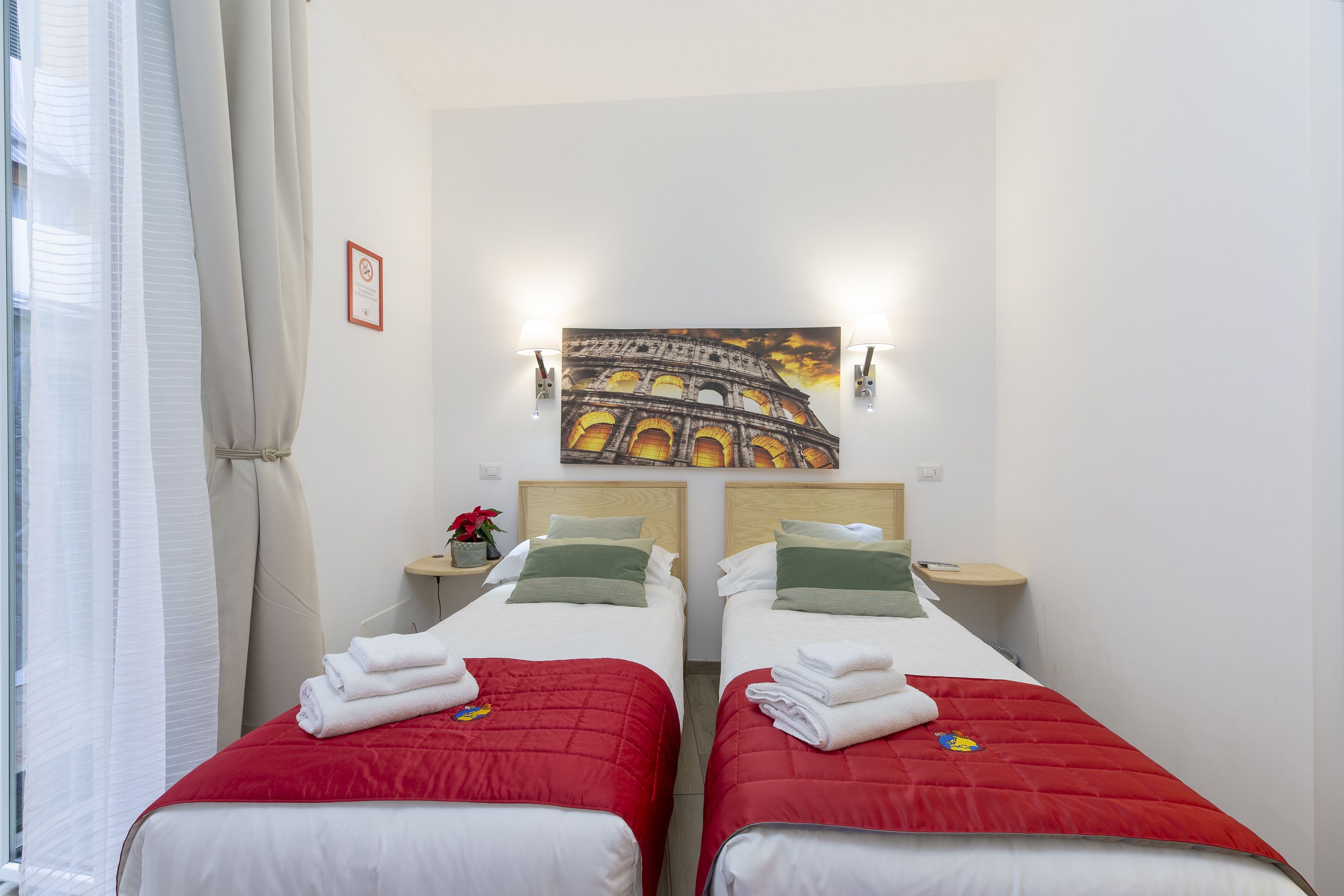 Een hotelkamer met twee bedden, elk voorzien van een rode sprei en witte lakens, een schilderij van het Colosseum aan de muur, en een klein tafeltje met een plant.