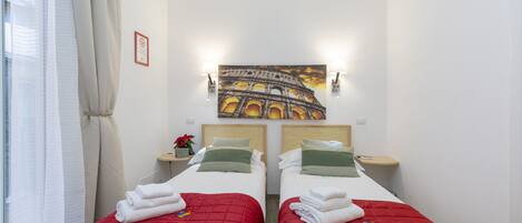 Een hotelkamer met twee bedden, elk voorzien van een rode sprei en witte lakens, een schilderij van het Colosseum aan de muur, en een klein tafeltje met een plant.