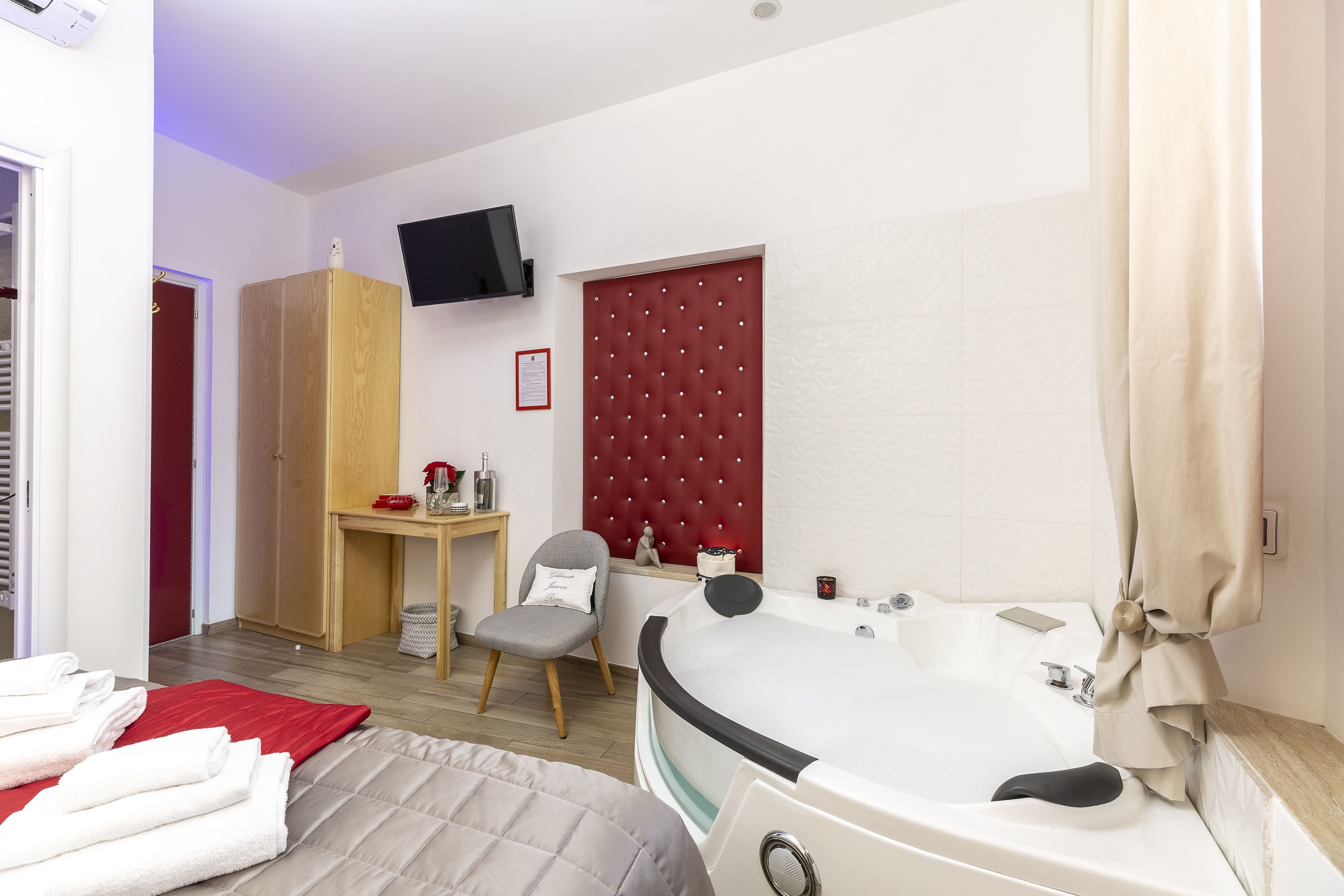 A hotel room with a large bed, a red headboard, a wooden wardrobe, a small table with a chair, a TV mounted on the wall, and a bathtub with a curtain.